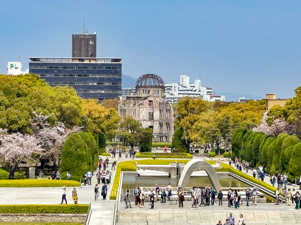 An aerial view of the peace park shows a monument standing in front of a pond with the atomic bomb dome in the background.