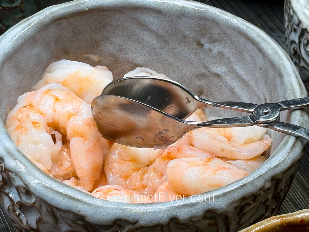 A large metal serving bowl contains large, cooked, and deshelled shrimps, accompanied by a pair of tomgs.