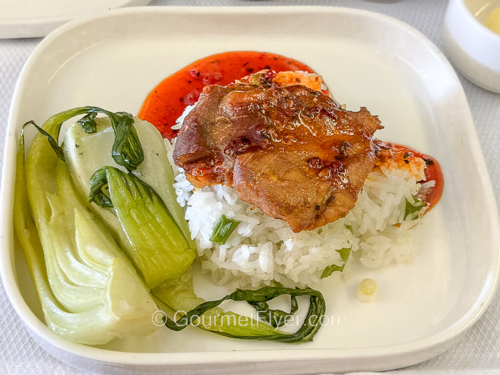 A piece of meat coated with a red sauce is served atop a bed of white rice and accompanied by Chinese vegetables.