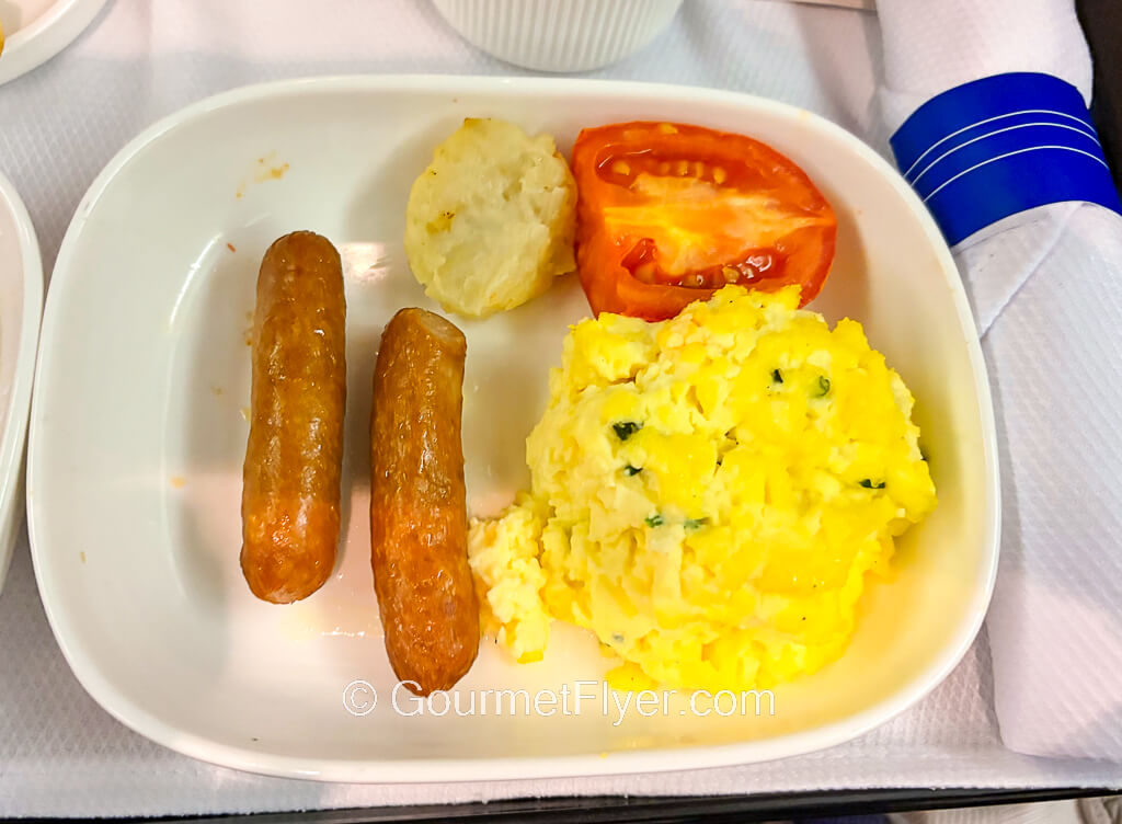 Scrambled eggs and a pair of sausages are accompanied by potatoes and a tomato wedge.