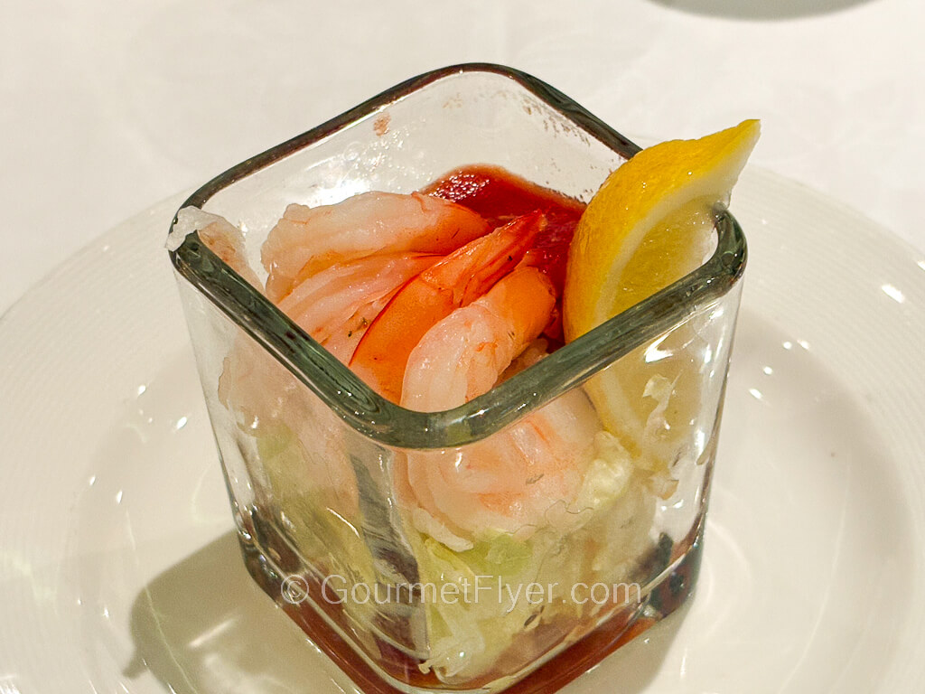 A glass contains four deshelled cooked shrimps served with a lemon wedge.