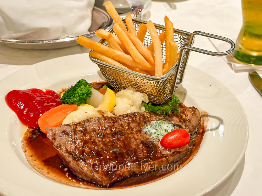 A steak is accompanied by a mini basket of French fries and vegetables.
