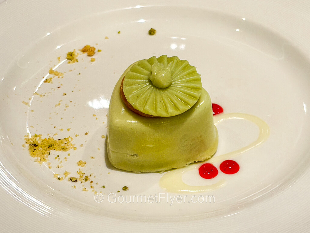 A greenish color mousse is decorated with drops of red syrups and cream on a plate.
