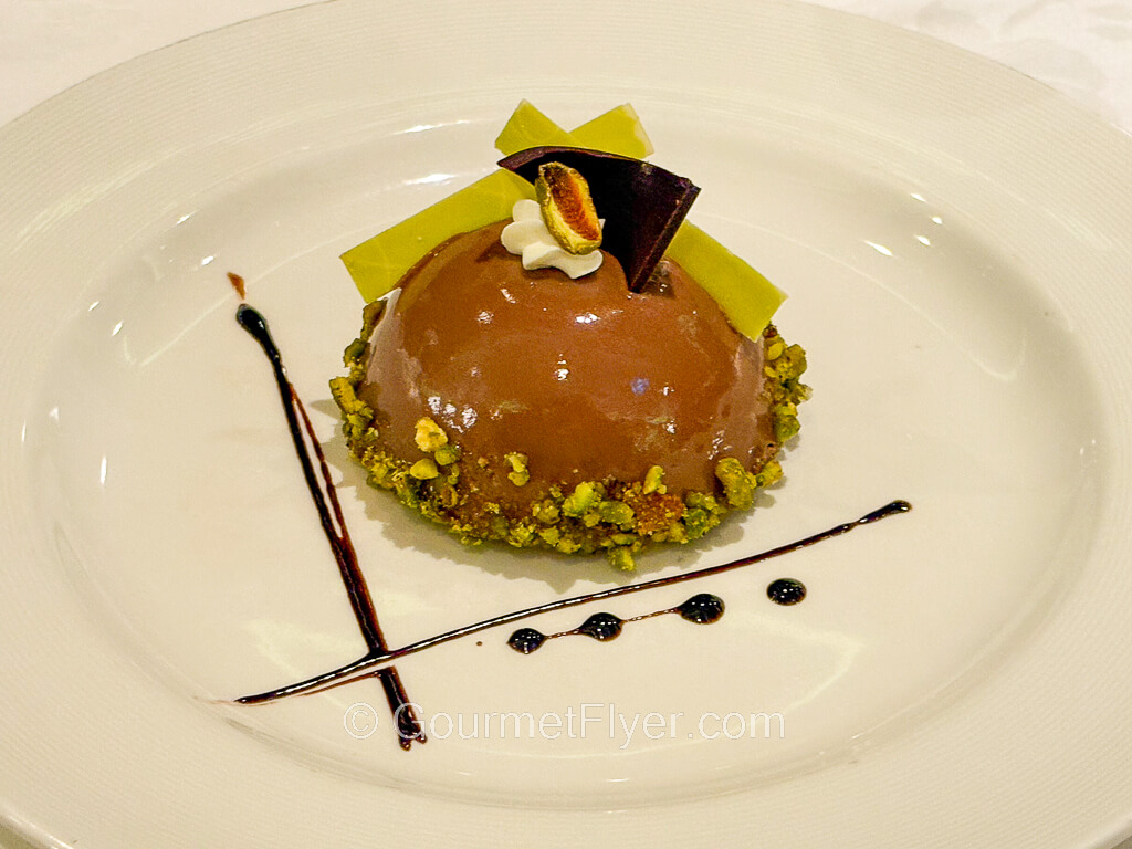 A dome-shaped chocolate dessert is garnished with chopped pistachios and decorated with chocolate syrup.