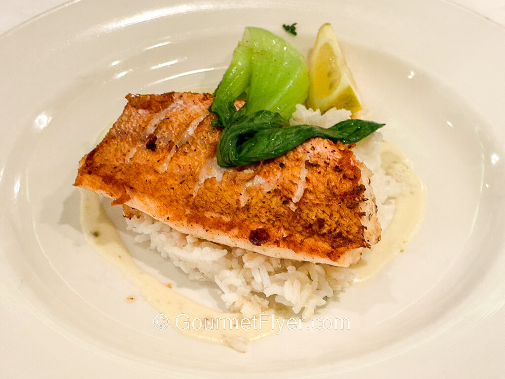A piece of grilled fish sits atop a bed of white rice and is accompanied by bok choy.