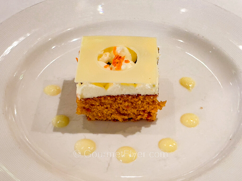 A square serving of a cake is garnished with droplets of cream around the plate.