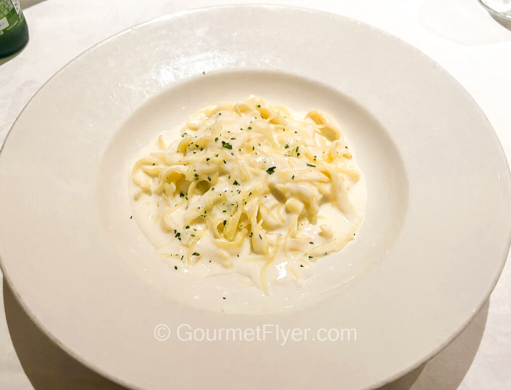 A plate of wide pasta is drenched in a creamy white sauce and garnished with parsley flakes.