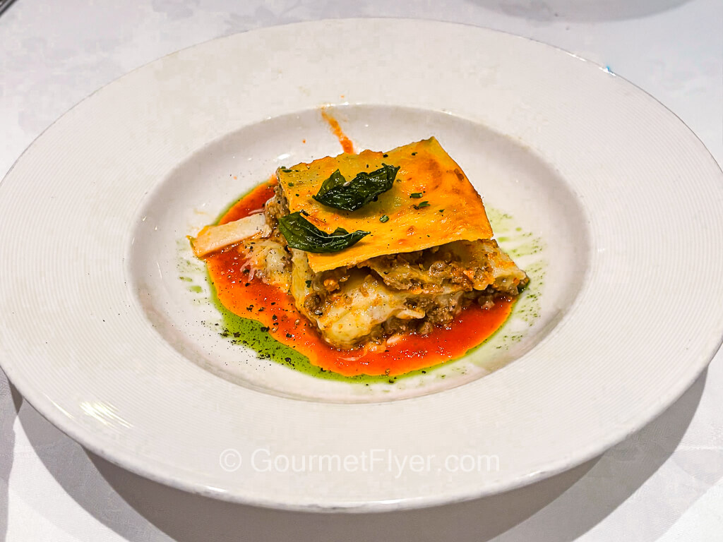 A square piece of lasagna is served in a shallow dish and garnished with red sauce and mint leaves.