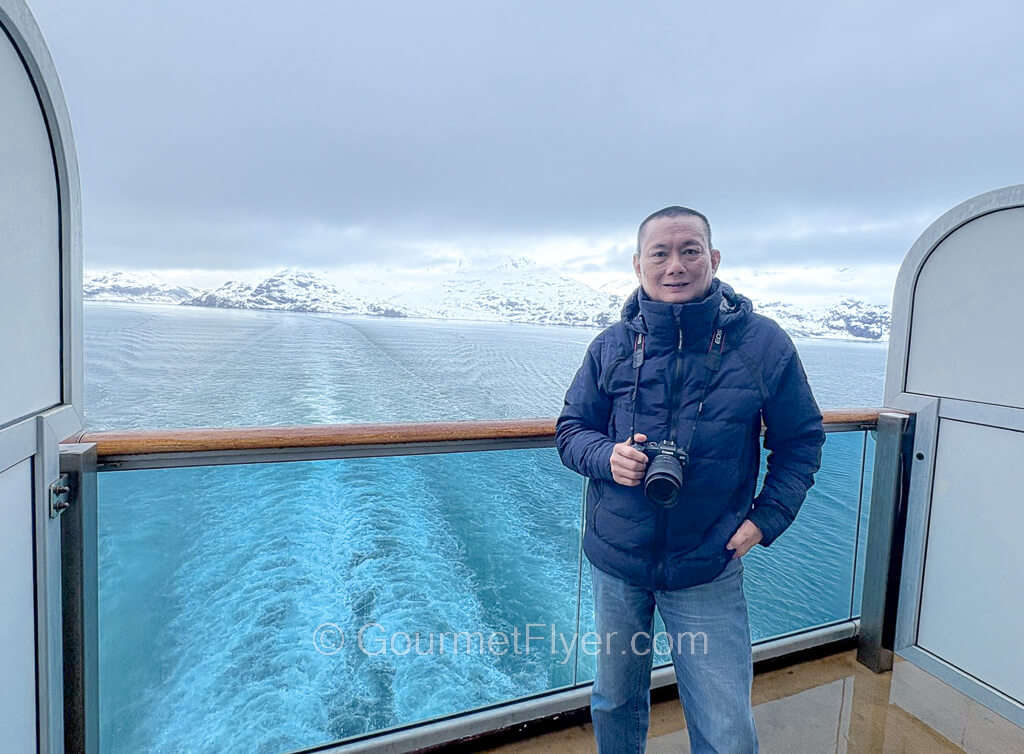 The blogger Gourmet Flyer poses in an aft-facing balcony with wake views and icy mountains in the background.