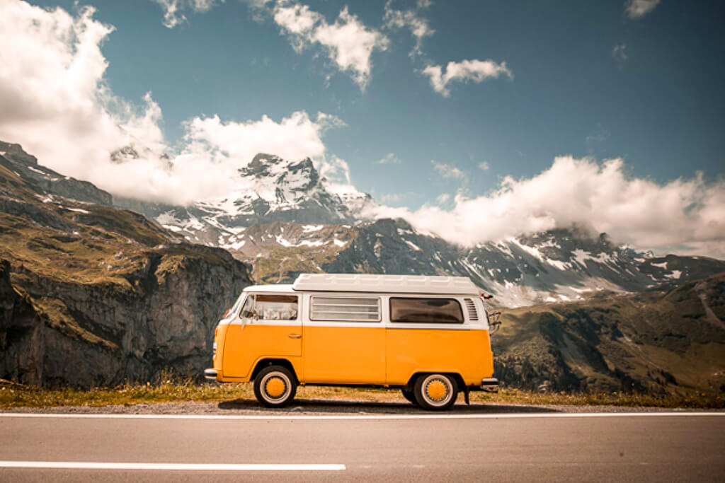 A yellow camper van is traveling under a blue sky with mountains in the backdrop.