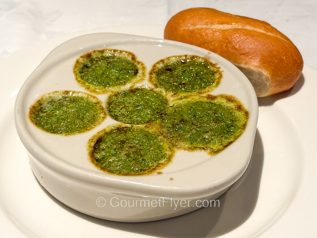A plate of snails in a green sauce is accompanied by a baquette.