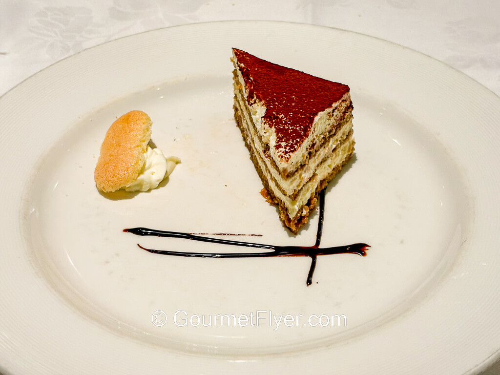 A slice of Tiramisu cake is garnished with chocolate syrup.