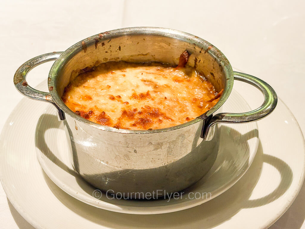 A stainless-steel bowl contains a soup with melted cheese on top.