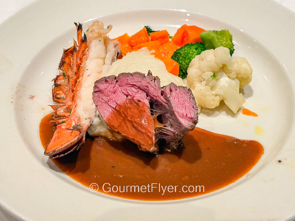 A medium rare beef filet is served with a split lobster tail and accompanied with vegetables.