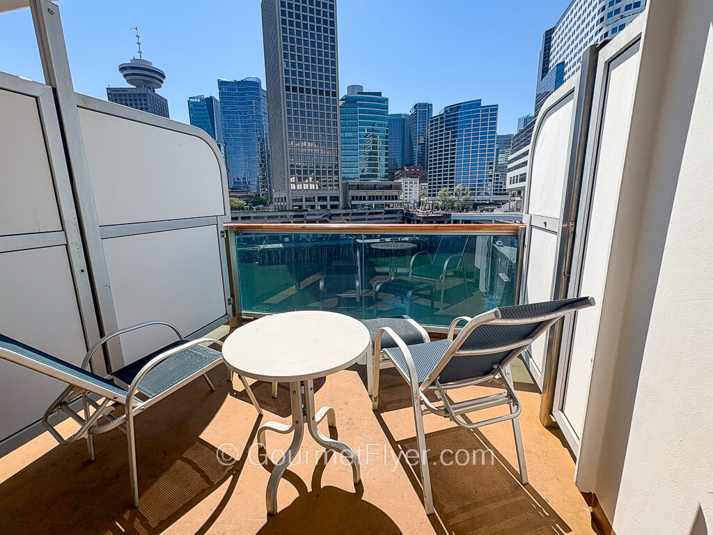 A cruise ship balcony with two lounge chairs and a small table faces the skyscrapers of a city.