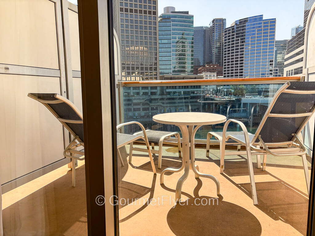 A closer view of a cruise ship balcony with two lounge chairs and a small table facing the skyscrapers of a city.