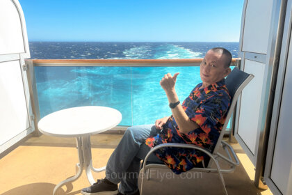 Review of Princess Royal Class Aft Facing Premium Deluxe Balcony features the blogger Gourmet Flyer relaxing on a deck chair in an aft balcony with wake view.
