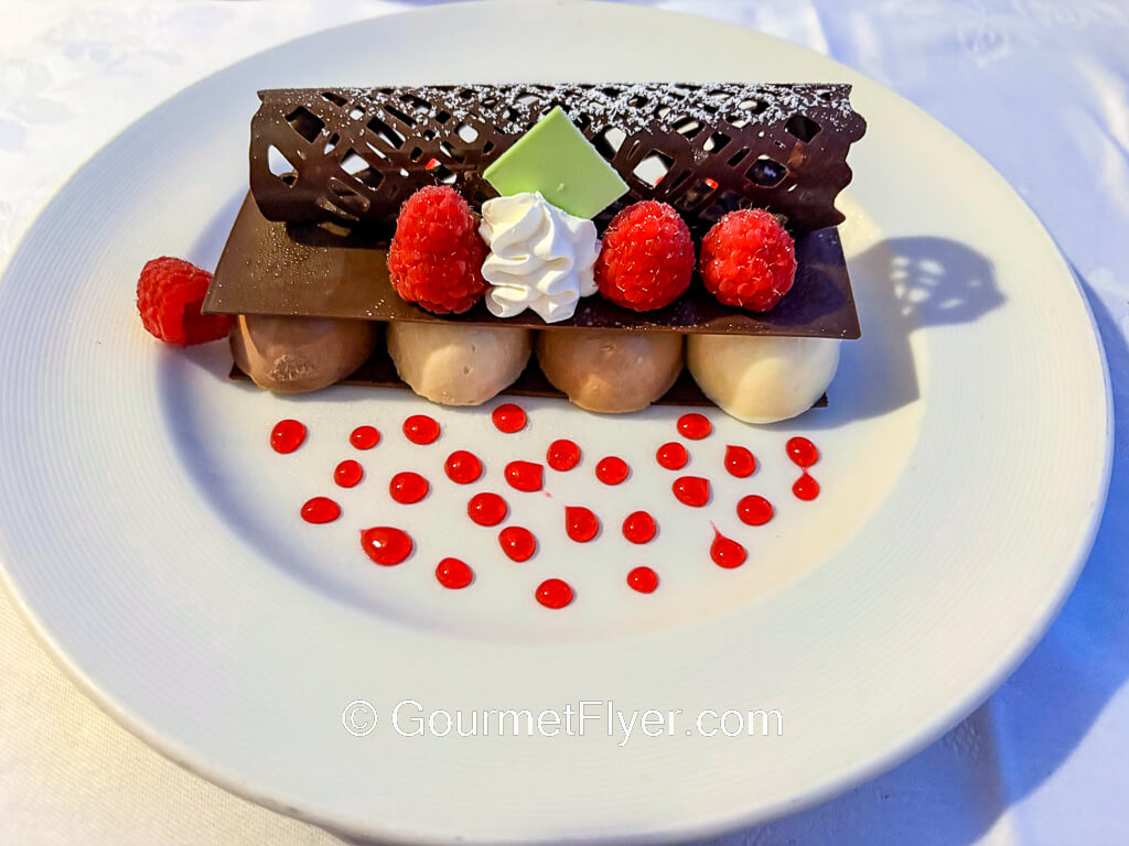 Pieces of mousse like desserts are garnished with berries, chocolate, and drizzles of red syrup.