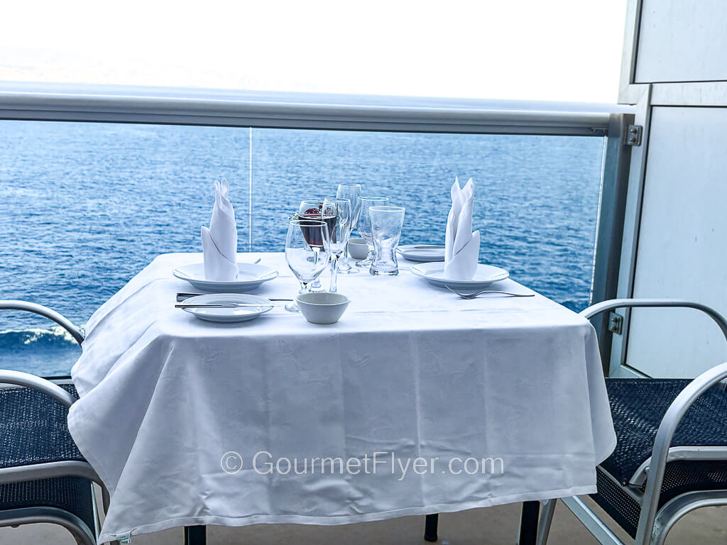 A dining table with white tablecloth and tableware is set up on a cruise ship balcony facing the ocean.
