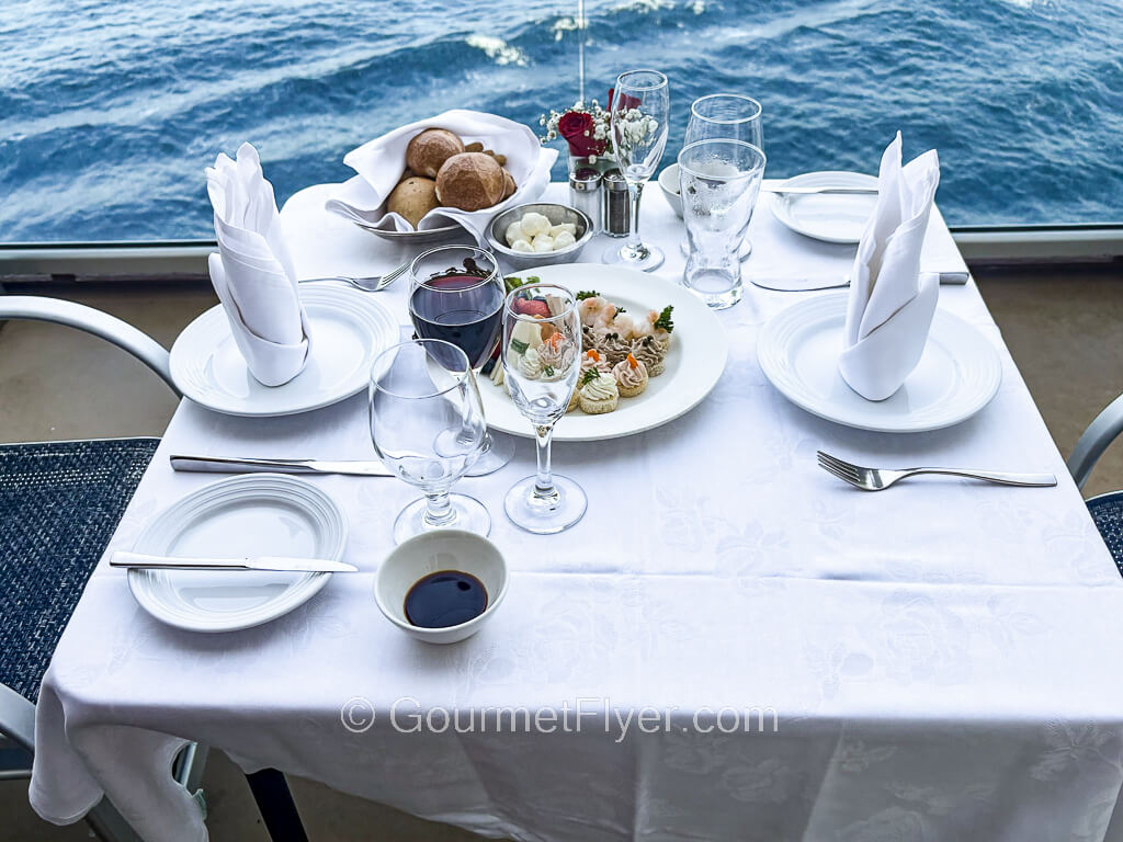 A tabletop with white tablecloth is set up with glassware, silverware and plates of food and drinks.