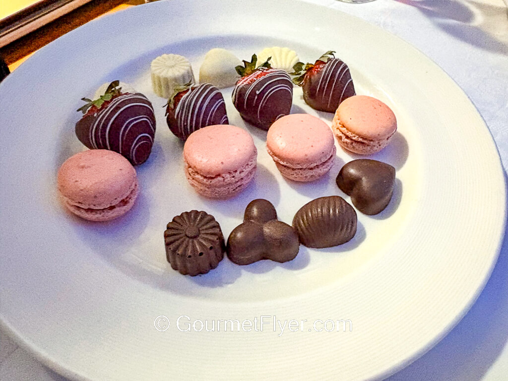 A plate is filled with chocolates, macarons, and chocolate coated strawberries.