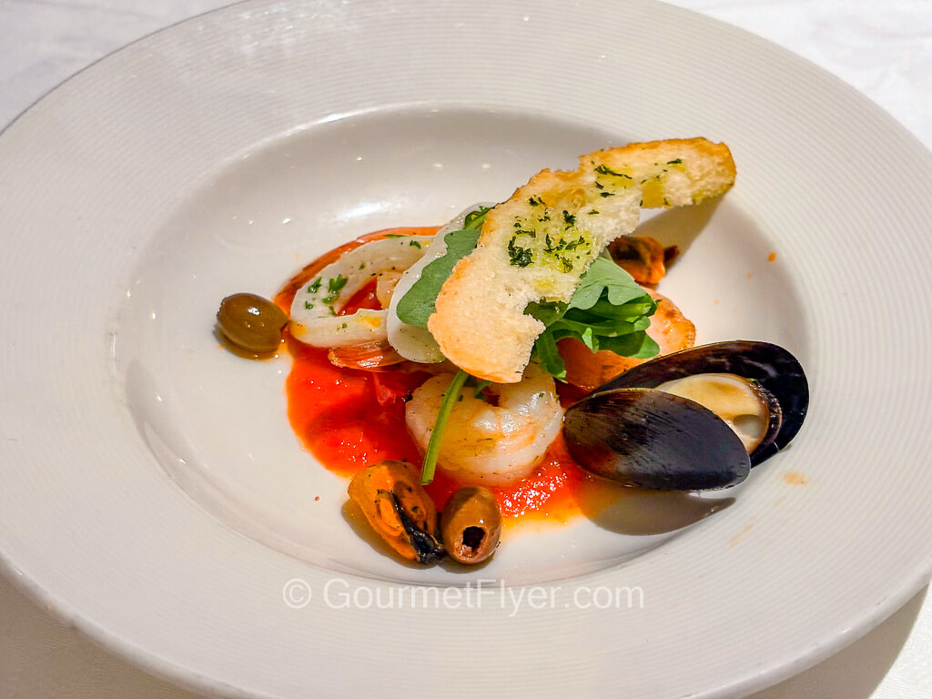 A plate of mixed seafood is served with a red sauce and accompanied with a slice of banquette.