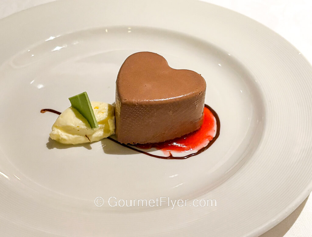 A heart-shaped chocolate dessert is garnished with a red sauce and whipped cream.