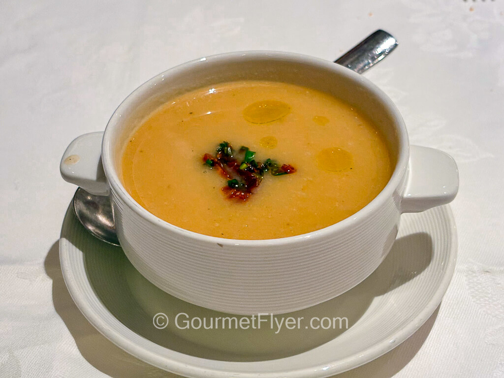 A orange colored soup is served in a bowl with garnishers on top.