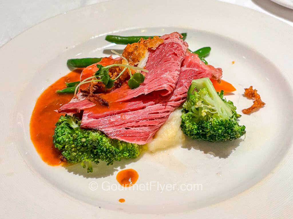 Slices of red deli meat is served with a brown gravy and accompanied with broccoli and mashed potatoes.